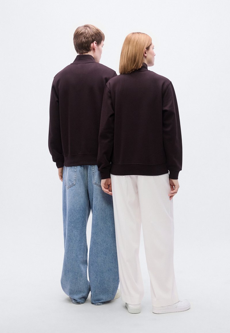 Deux personnes sont debout dos à dos, portant un sweat-shirt marron foncé et un pantalon large de couleur claire. Le tissu semble doux avec une coupe décontractée.
