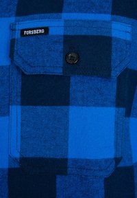 Camisa de cuadros azul con cuadrados en tonos azules más oscuros y más claros, un bolsillo en el pecho con botón y una pequeña etiqueta "Forsberg". Textura suave.