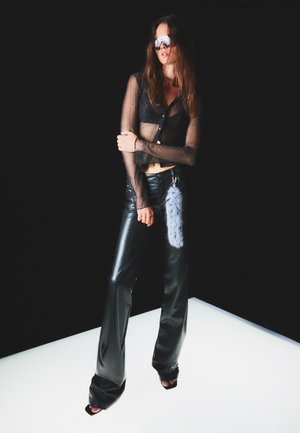 Schwarze, aus Kunstleder gefertigte weite Hosen kombiniert mit einem transparenten, schwarzen Langarmoberteil mit Knöpfen. Als Accessoire dient ein graues Fell-Charm.