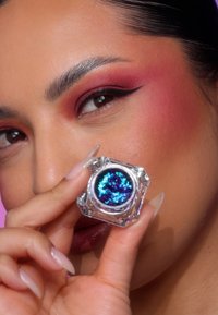 En sekskantet klar beholder som holder et iriserende blått og lilla kosmetisk produkt. Nærbilde av en hånd med lange, lyse negler og fargerik øyemakeup.