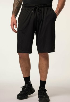 Mand iført sorte shorts med snøre, sorte sportssko og sorte sokker, står med afslappede hænder og den ene hånd i lommen.