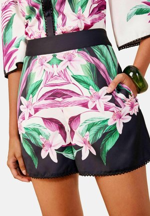 Kvinde iført shorts med blomsterprint og matchende top med grønne og lyserøde blomster, sort taljebånd og et grønt armbånd på håndleddet.