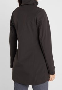 Chaqueta negra de material suave con un alto cuello, mangas largas y corte recto. Presenta una ventilación trasera y un diseño minimalista.