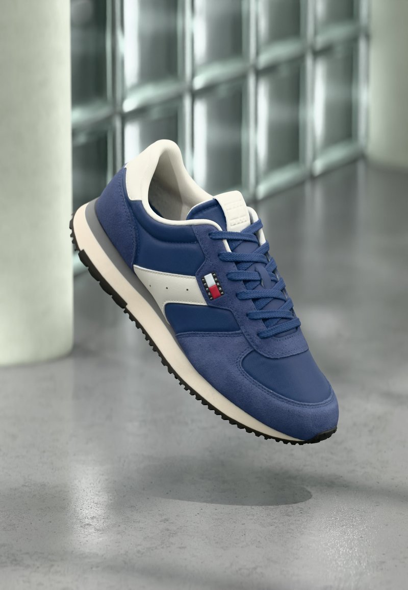 Baskets bleu et blanc avec semelle noire, logo carré rouge-blanc-bleu sur le côté, présentées sur un sol gris avec un mur en blocs de verre en arrière-plan.