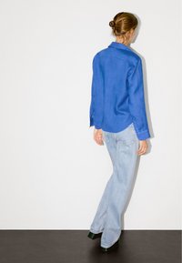 Hellblaue Leinenbluse mit Kragen und knöpfigem Ärmelsaum, kombiniert mit hellblauen, weit geschnittenen Jeans und schwarzen Plateau-Schuhen, von hinten betrachtet.