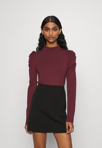 Burgundy long-sleeve top with puffed shoulders, paired with a black mini skirt, smooth fabric, fitted silhouette, and high neckline.