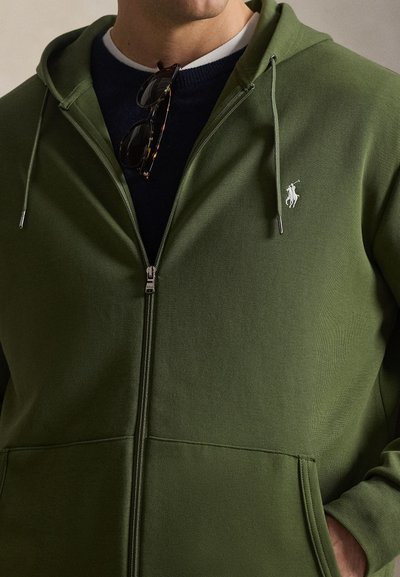 Mees kannab oliivrohelise lukuga dressipluusi, millel on valge Polo Ralph Laureni logo rinnal, ning päikeseprille, mis ripuvad mereväe sinise särkkaela küljes.