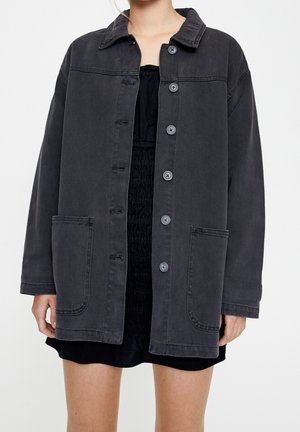 Vrouw draagt een oversized donkergrijze jas met grote knopen en grote voorzakken, over een zwarte jurk met plooien, tegen een effen achtergrond.