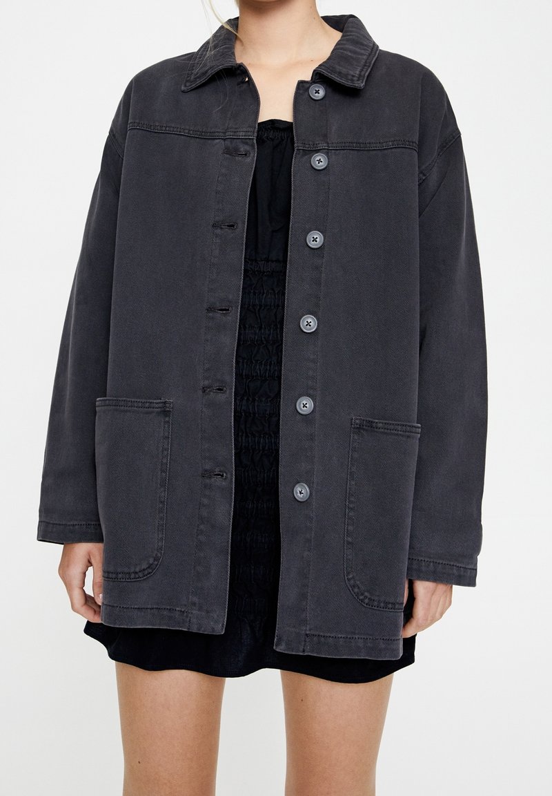 Femme portant une veste ample gris foncé à boutons avec de grandes poches frontales, par-dessus une robe noire froncée, sur un fond uni.