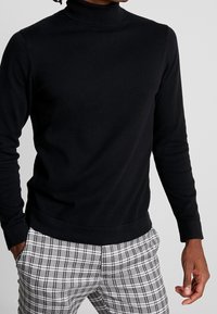 Suéter de cuello alto negro en tejido de punto suave con mangas largas, combinado con pantalones de cuadros gris claro con detalles en negro y ajuste slim.
