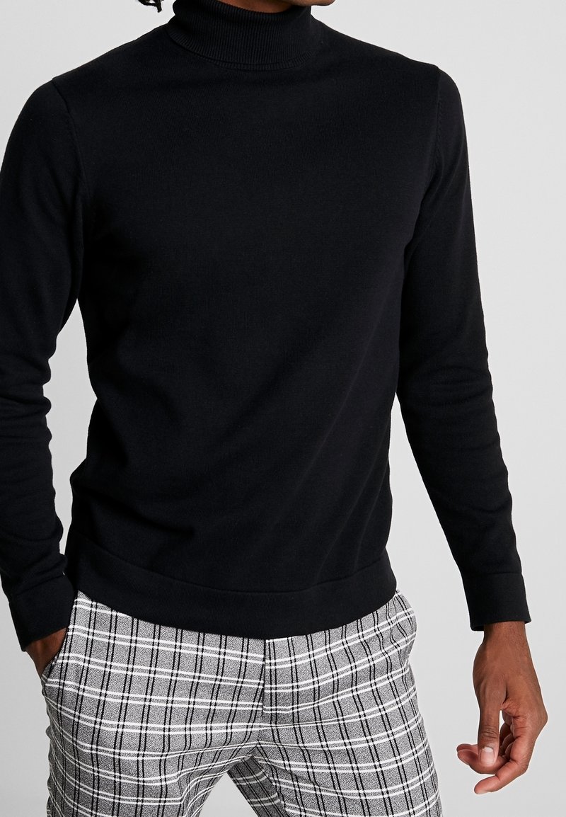 Suéter de cuello alto negro en tejido de punto suave con mangas largas, combinado con pantalones de cuadros gris claro con detalles en negro y ajuste slim.