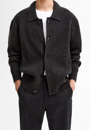 Cardigan côtelé gris foncé avec un col, des boutons à l'avant et des manches longues, porté sur une chemise blanche et associé à un pantalon sombre.