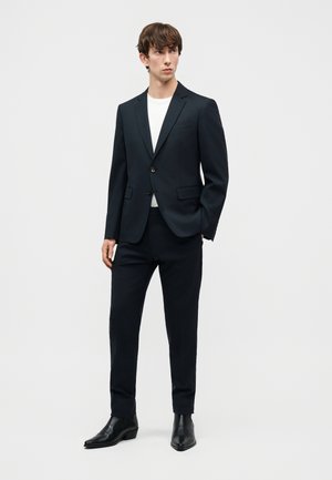 Νεαρός άνδρας στέκεται, φορώντας μαύρο κοστούμι με λευκό πουκάμισο και μαύρα δερμάτινα μποτάκια, μπροστά σε απλό ανοιχτό φόντο.