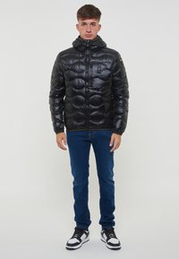 Giacca a vento nera con cappuccio, design trapuntato, zip frontale e patch con logo, abbinata a jeans blu e sneakers nere e bianche.