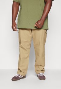 Beige cargobroek met zijzakken, losse pasvorm en regulier stiksel. Gecombineerd met donkerpaarse sneakers met witte strepen.