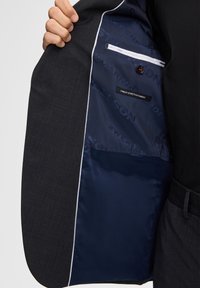 Donkergrijze blazer met een marineblauwe satijnen voering. Kenmerkt zich door een witte bies, een klein zakje en een etiket met de tekst "Finest Stretch Fabric."