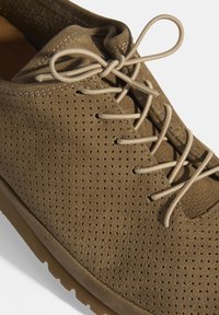 Chaussure en suede marron avec une texture perforée, dotée de lacets beige clair et d'une semelle en caoutchouc, présentant une forme de bout arrondi.