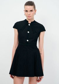 Schwarzes Spitzenkleid mit hohem Ausschnitt, kurzen Puffärmeln, zentralen Knöpfen und einem ausgestellten Rock. Mit einem Stoffgürtel und floralen Knopfakzenten.