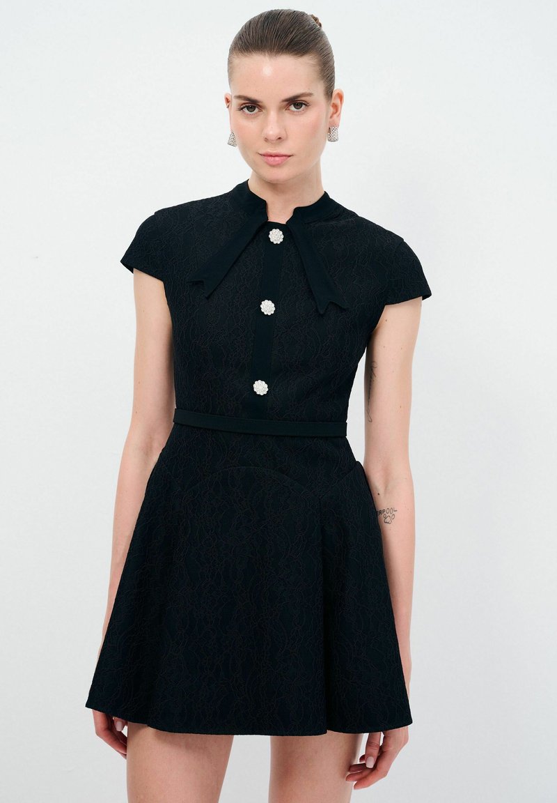 Schwarzes Spitzenkleid mit hohem Ausschnitt, kurzen Puffärmeln, zentralen Knöpfen und einem ausgestellten Rock. Mit einem Stoffgürtel und floralen Knopfakzenten.