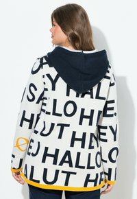 Femme aux cheveux bruns portant un sweat à capuche blanc avec un texte de salutations noir et une capuche bleu marine, se tenant devant un fond blanc uni.
