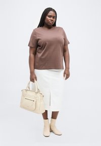 Γυναίκα με μακριά πλεξούδα, φοράει καφέ μπλουζάκι, λευκή φούστα, μπεζ μποτάκια στον αστράγαλο και κρατάει μια μπεζ τσάντα στο χέρι, με φόντο λευκό.