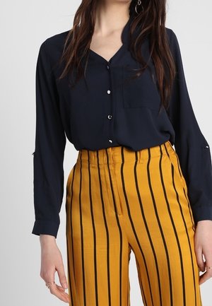 Kobieta w granatowej bluzce na guziki, wsuniętej w musztardowożółte spodnie z pionowymi czarnymi paskami, widoczne jej tułów i dłonie.