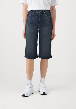 Shorts en denim bleu foncé, avec une coupe décontractée et des bords retroussés. Portés avec des chaussures de sport blanches et des chaussettes.