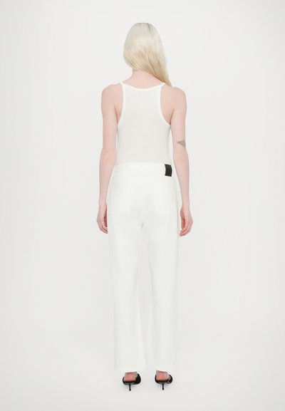 Pantalons larges blancs avec taille haute, présentant une découpe au dos et un détail de tag noir. Portés avec un débardeur blanc ajusté.