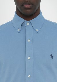 Cămașă bleu deschis, cu butoni, confecționată dintr-un material texturat. Dispune de un guler clasic, butoni albi și un mic logo închis la culoare pe piept.