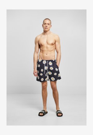 Ung mand står barfodet iført sorte badeshorts med hvide og gule margeritprints samt sorte slidesandaler med hvid tekst.