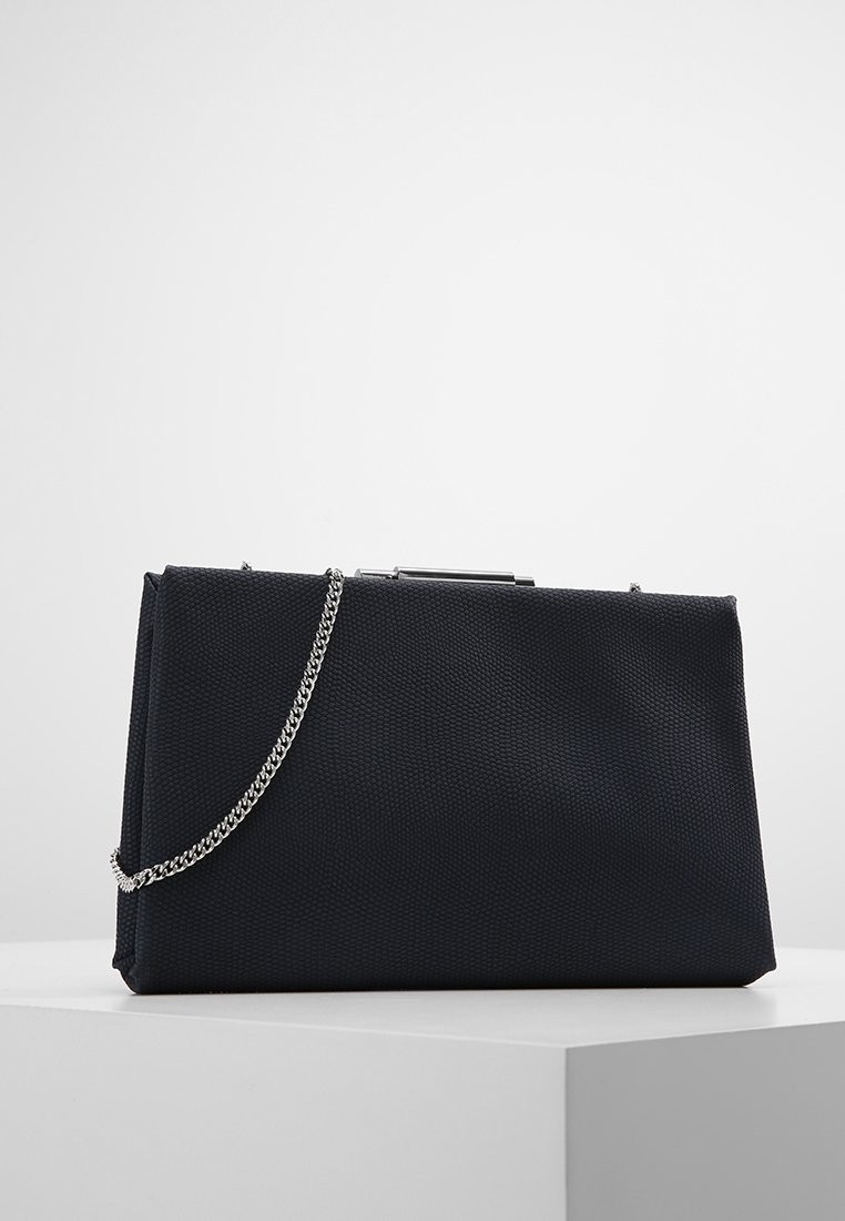 Pochette noire texturée avec une bandoulière chaîne argentée et un fermoir métallique, exposée sur un socle blanc sur un fond gris clair uni.