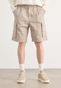 Calções de carga bejes com uma cintura elástica, dois bolsos laterais e um corte descontraído, combinados com sapatos bejes claros e meias brancas.