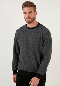 Hallikas tekstuuriga dressipluus pikkade varrukatega, musta sooniku kaeluse ja mansettidega ning sirge alläärega. Esivaat, seljas meesmudel.