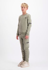 Blonde jongen staat in een olijfgroene sweatshirt en bijpassende cargopants met beige sneakers tegen een witte achtergrond.