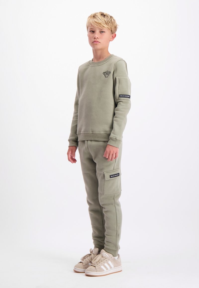 Blonde jongen staat in een olijfgroene sweatshirt en bijpassende cargopants met beige sneakers tegen een witte achtergrond.