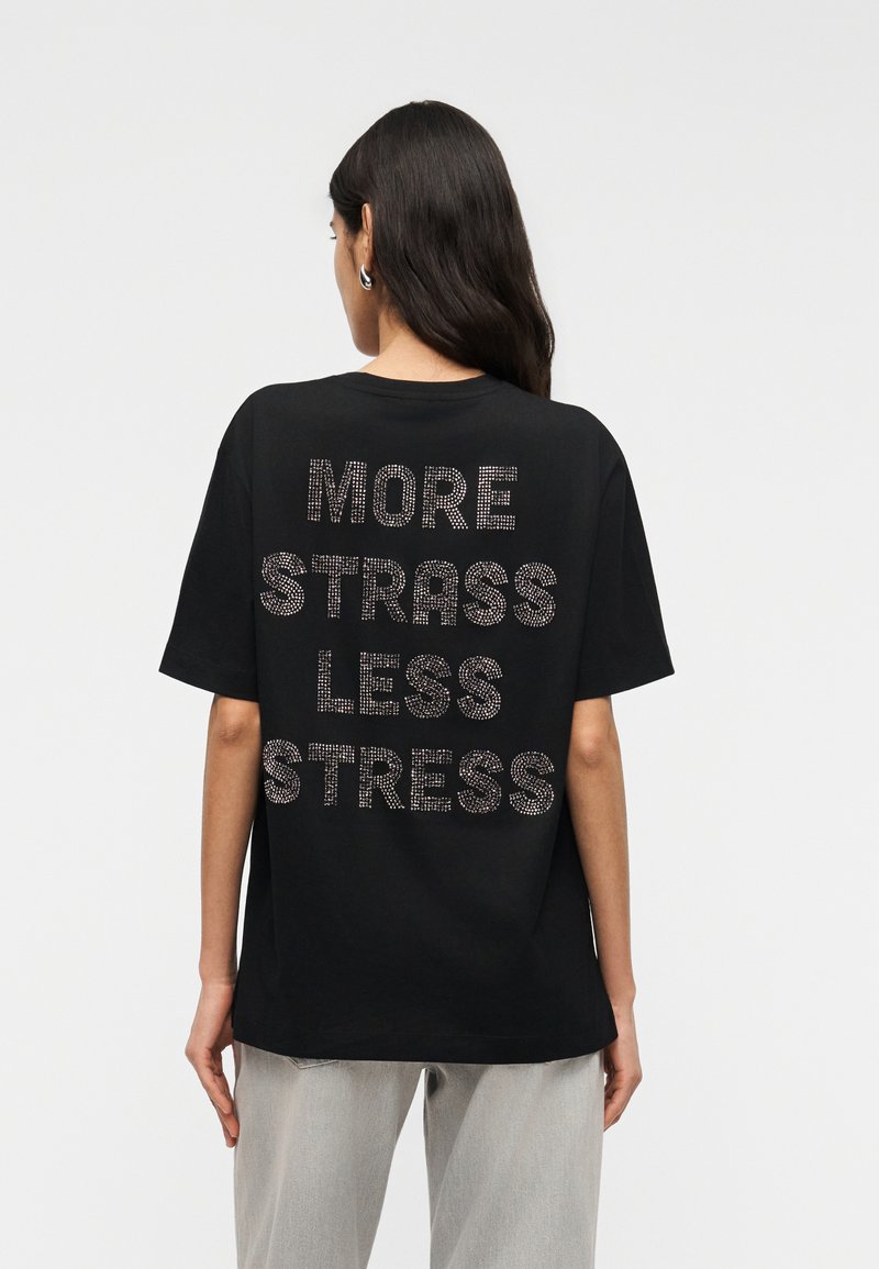 Γυναίκα με μακριά σκουρόχρωμα μαλλιά, που φοράει μαύρο t-shirt με ασημένια καρφιά που σχηματίζουν τη φράση «MORE STRASS LESS STRESS» στην πλάτη, συνδυασμένο με ανοιχτό γκρι παντελόνι.