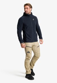 Marineblauer Fleece-Reißverschluss-Hoodie mit hohem Kragen, kombiniert mit beigen Cargo-Hosen mit Reißverschlusstaschen und einem schwarzen Logoaufnäher. Schwarze Schuhe.