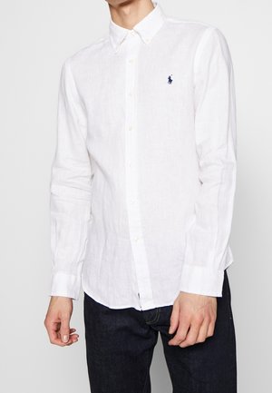 Mann trägt ein weißes langärmliges Hemd mit blauem gesticktem Logo auf der Brust und dunkle Jeans.