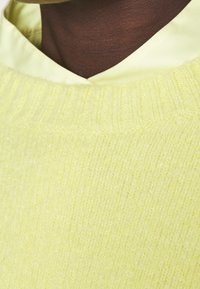 Hellgelber Strickpullover mit einem gerippten Ausschnitt, der eine glatte Textur aufweist und über einem blassgelben Hemd mit Kragen getragen wird.