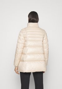 Kobieta z długimi ciemnymi włosami, ubrana w beżową kurtkę puchową i czarne spodnie, widziana z tyłu na jednolitym białym tle.