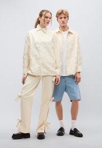 Δύο μοντέλα φορούν ανοιχτόχρωμα ρούχα. Η γυναίκα έχει μια ριγωτή μπεζ μπλούζα και παντελόνια που ταιριάζουν με φιόγκους. Ο άνδρας φορά ένα ανοιχτόχρωμο πουκάμισο και σορτς από denim.