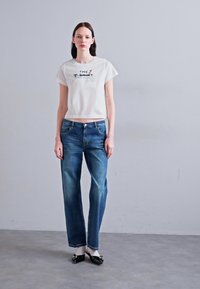 Hvit, kort t-skjorte med grafisk tekst, kombinert med løstsittende blå denimjeans. Modellen har på seg svarte leppestifter. Enkel, uformell antrekk.