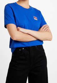 Blå bomullströja med rund halsringning och korta ärmar, som har en liten röd och vit logotyp på vänster bröst. Kombineras med svarta jeans.