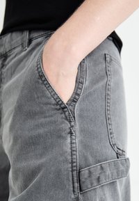 Grå denimbukser har en sidelomme med en rund åpning, synlig søm og en teksturert overflate. Designet er avslappet og funksjonelt.