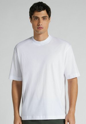 Ung mand med mørkt hår iført en simpel hvid T-shirt med korte ærmer og rund hals, stående mod en grå baggrund.