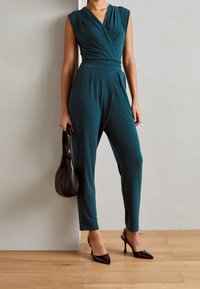 Mujer vestida con un mono sin mangas de color verde azulado oscuro, tacones negros de punta, sosteniendo un bolso marrón oscuro, de pie sobre un suelo de madera junto a la pared.