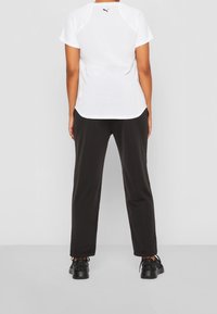 Valkoinen lyhythihainen urheilupaita, jossa musta logo takana, yhdistettynä mustiin kapeneviin housuihin ja mustiin lenkkareihin, näkymä takaa.