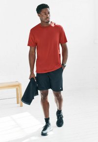 Rotes, kurzärmeliges Sportshirt, schwarze Shorts und schwarze Sportschuhe; hält ein dunkles Handtuch. Glatte Stoffe und lockerer Schnitt. Minimales Design.