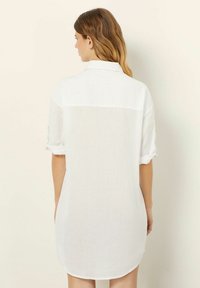 Abito camicia bianco in lino con colletto, maniche arrotolate e un orlo di lunghezza media. Presenta un tessuto morbido e testurizzato con fondo curvo.
