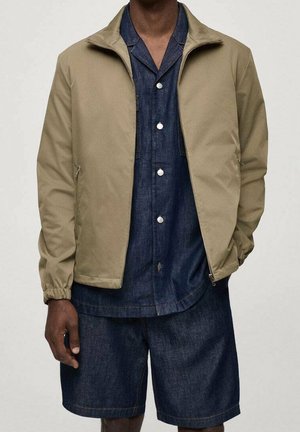 Mand iført beige lynlåsjakke over mørkeblå skjorte med knapper og matchende shorts, stående med den ene hånd i lommen.